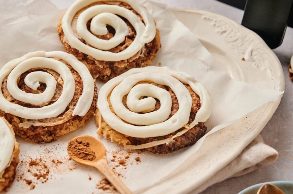 Three Air Fryer Crumbl Cinnamon Swirl Cookies with white icing spirals are arranged on parchment paper atop a white plate, with a dusting of cinnamon powder nearby.