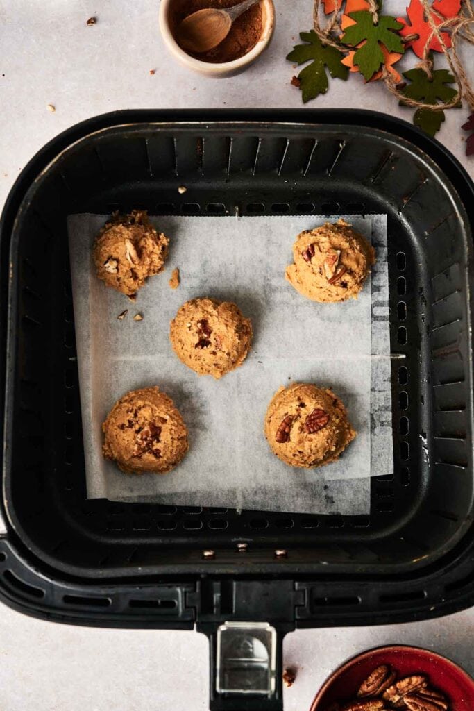 Five unbaked Air Fryer Pumpkin Cookies dough balls rest on parchment paper inside an air fryer basket, surrounded by fall-themed decor and small bowls.