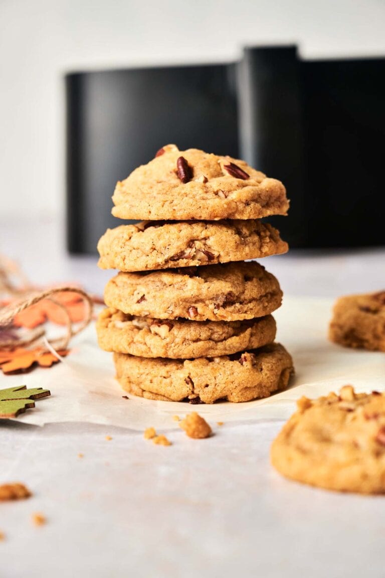 A stack of five Air Fryer Pumpkin Cookies on parchment paper with one cookie and crumbs nearby; blurred background features a black object.