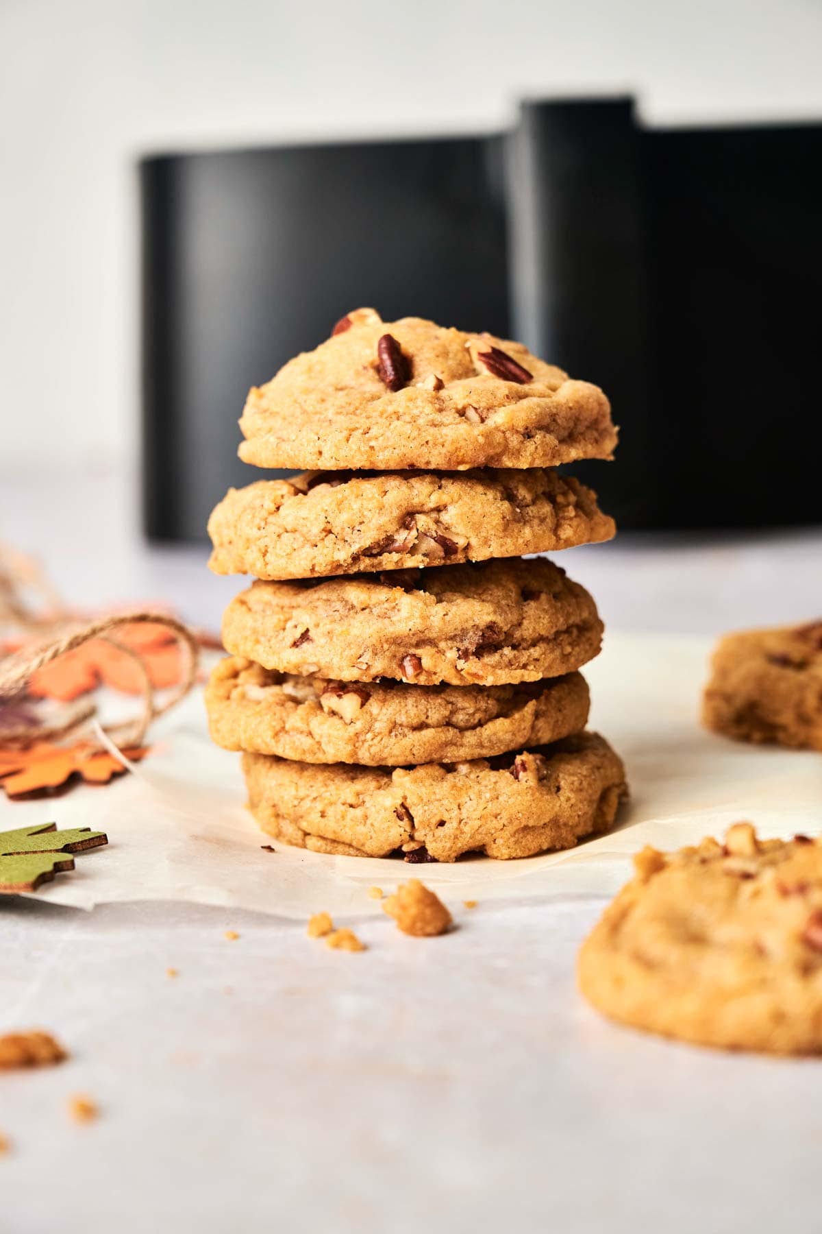 A stack of five Air Fryer Pumpkin Cookies on parchment paper with one cookie and crumbs nearby; blurred background features a black object.