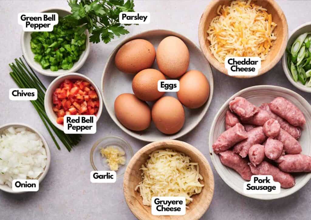 Various labeled bowls containing eggs, cheddar cheese, gruyere cheese, pork sausage, bell peppers, chives, onion, garlic, parsley, and sliced green onions&mdash;perfect ingredients for a delicious Breakfast Casserole&mdash;are arranged on the countertop.