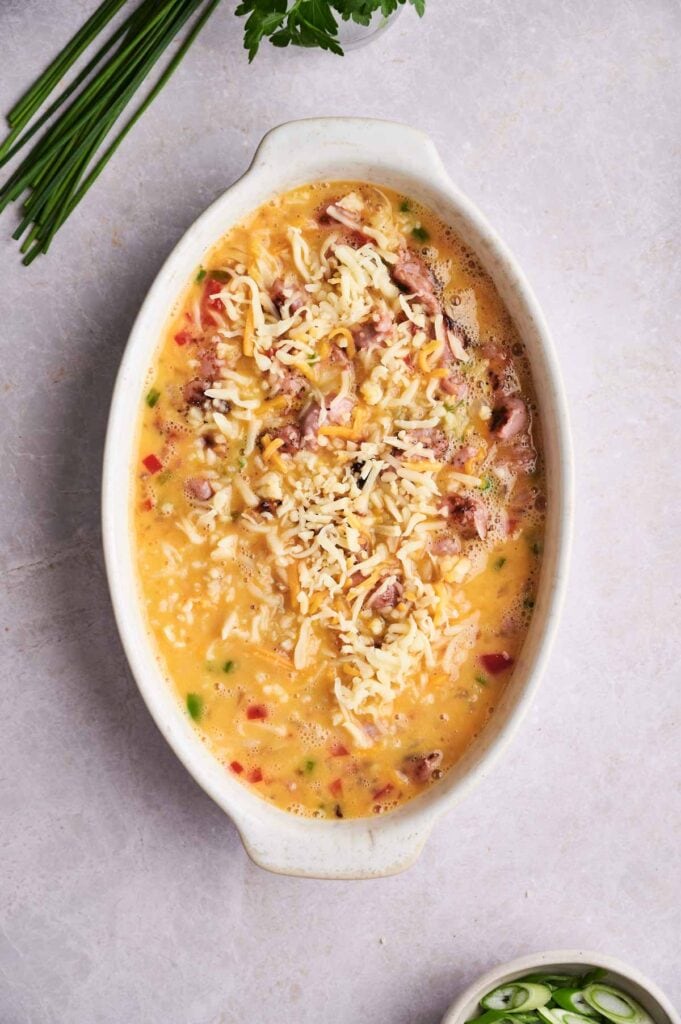 Oval baking dish filled with an uncooked egg mixture, diced vegetables, ham, and shredded cheese on a light countertop with herbs nearby&mdash;perfect for making a delicious Breakfast Casserole.