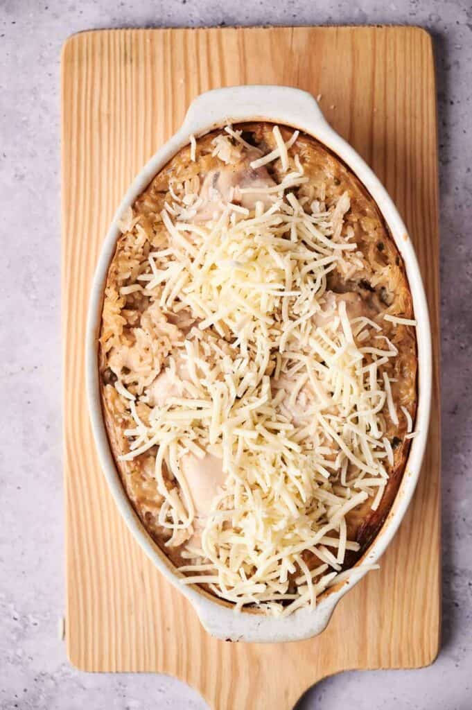 An oval white baking dish filled with a Chicken and Rice Casserole topped with shredded cheese, placed on a wooden cutting board.