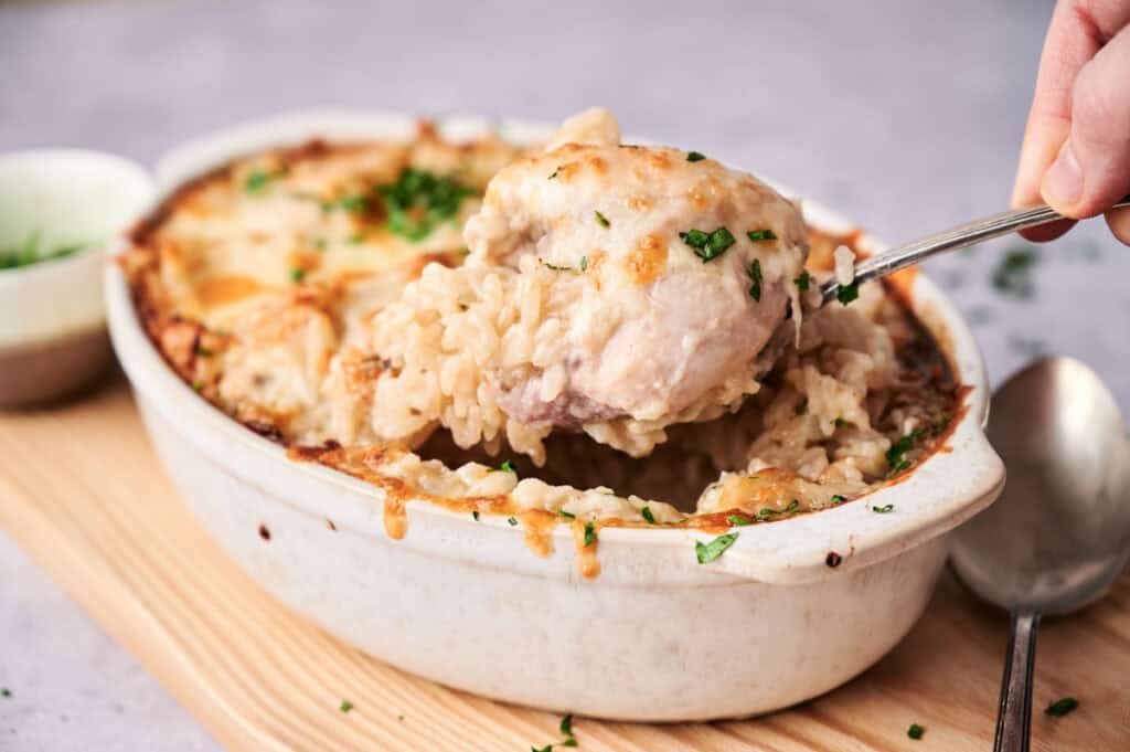 A hand serves Chicken and Rice Casserole from a white baking dish, topped with melted cheese and chopped herbs for a comforting meal.