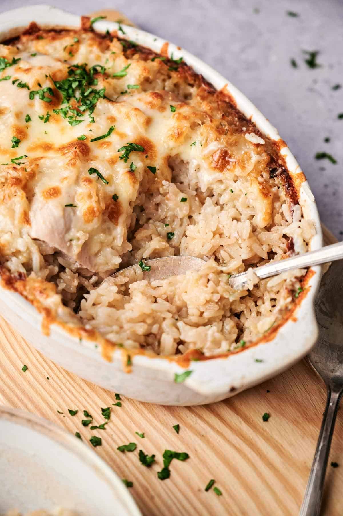 A Chicken and Rice Casserole dish filled with creamy rice and chicken, topped with melted cheese and garnished with chopped parsley, served hot with a spoon inside.