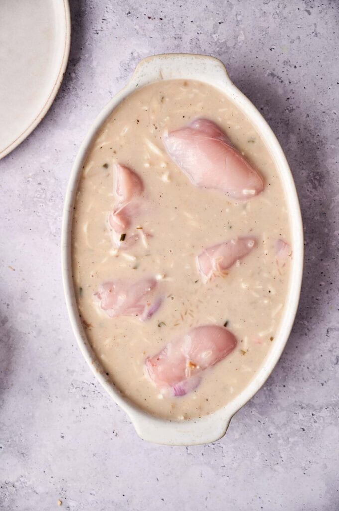 Raw chicken breasts in a creamy, seasoned mixture await baking for a classic Chicken and Rice Casserole, shown in an oval dish on a light textured surface.