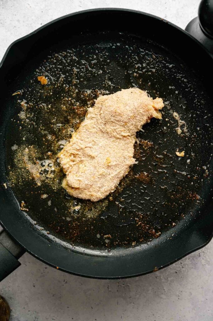 Chicken cutlets are being breaded and fried in oil in a black skillet.