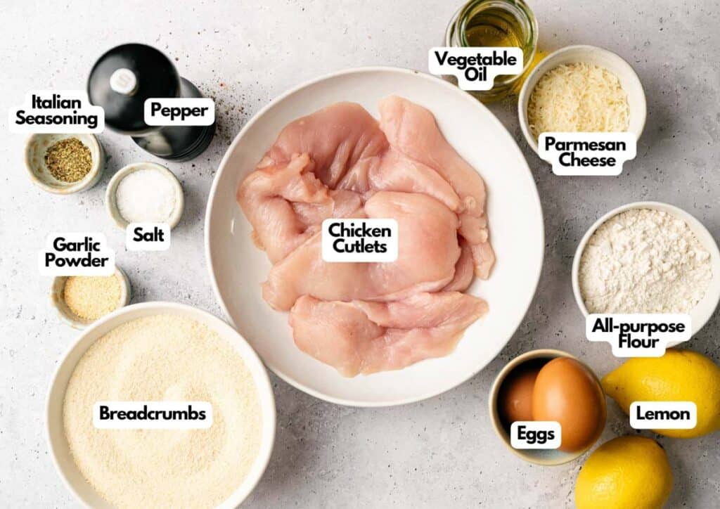 A bowl of raw chicken cutlets surrounded by ingredients including eggs, lemon, flour, parmesan cheese, breadcrumbs, vegetable oil, seasonings, salt, pepper, and garlic powder&mdash;perfect for making classic Chicken Cutlets.