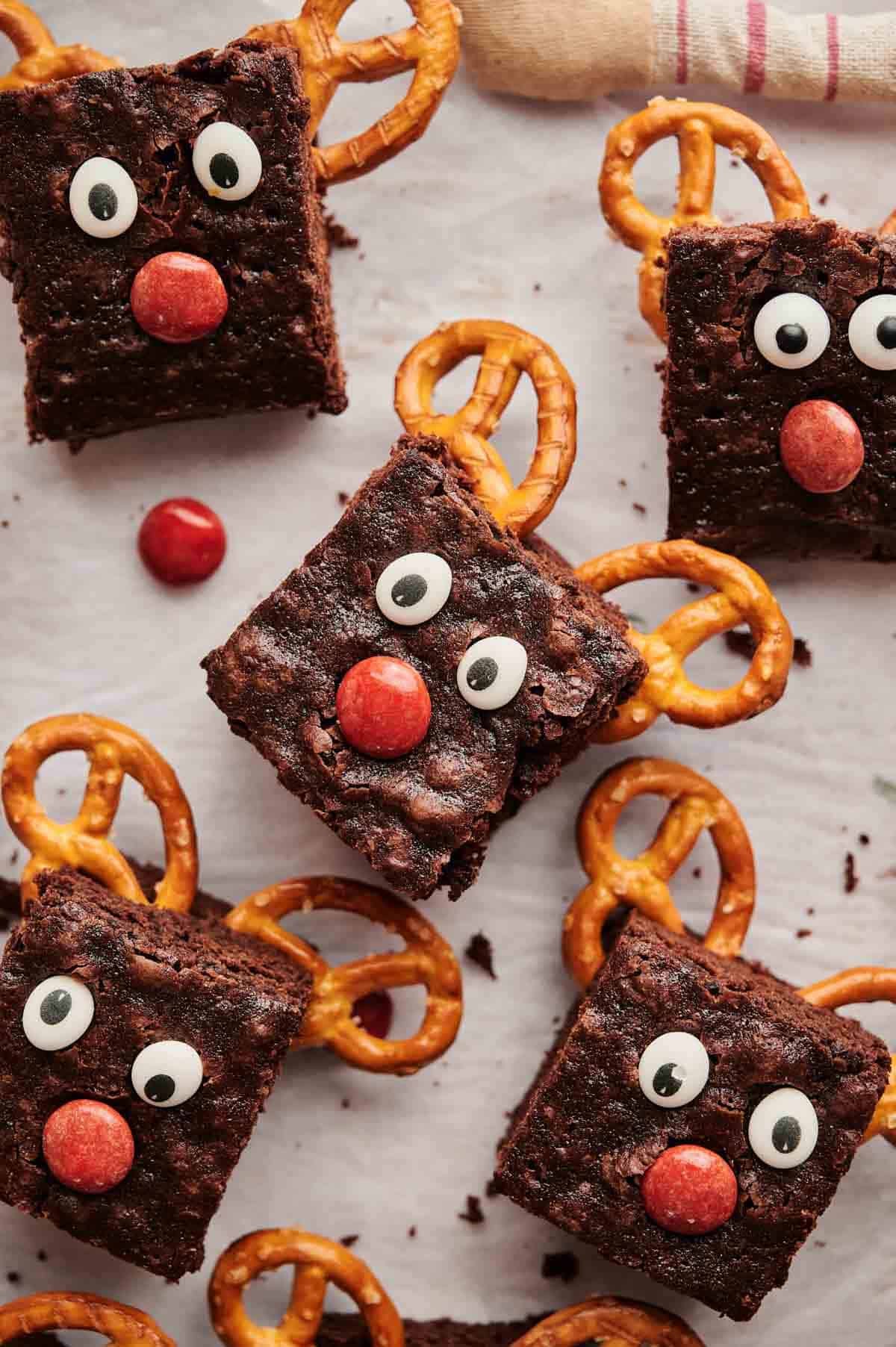 Christmas Brownies decorated as reindeer with pretzel antlers, candy eyes, and red noses are arranged on a white surface.