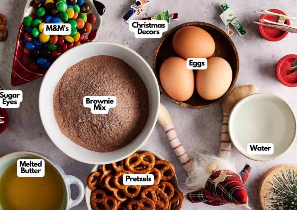 Top-down view of labeled ingredients for Christmas Brownies, including brownie mix, eggs, M&M's, festive decorations, sugar eyes, melted butter, pretzels, and water.