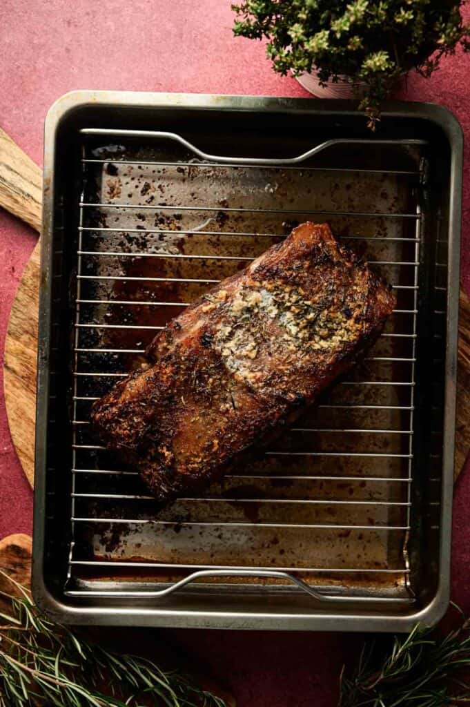 A Christmas Prime Rib Roast sits cooked on a wire rack in a roasting pan, surrounded by cutting boards and herbs on a festive red surface.