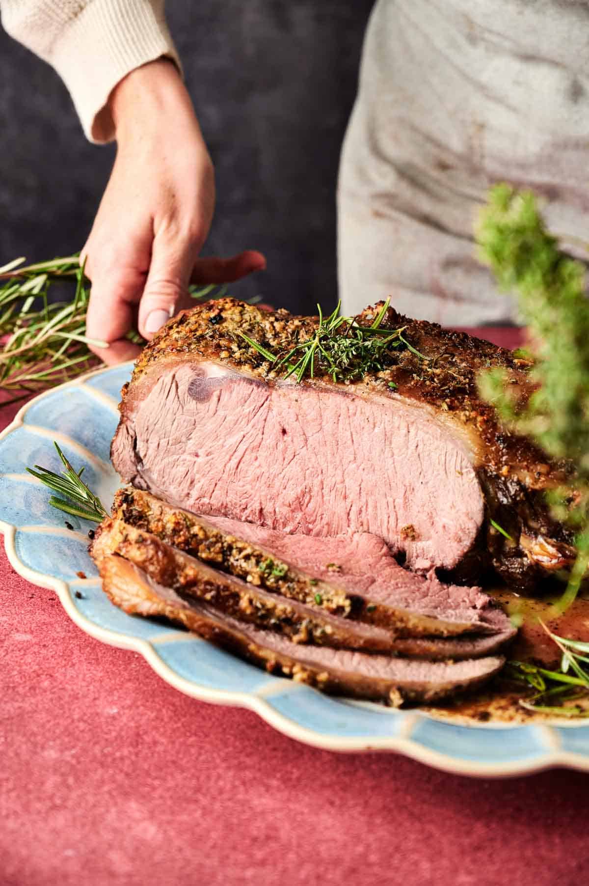 A person serves slices of Christmas Prime Rib Roast on a blue platter, garnished with fresh rosemary.