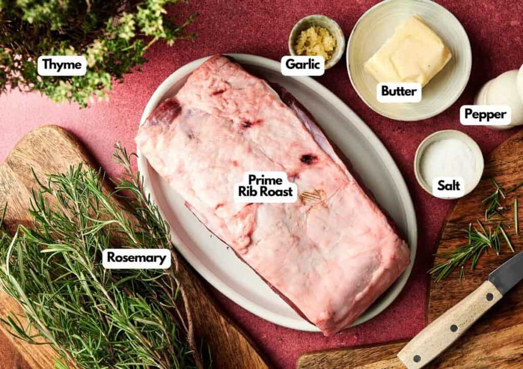 Christmas Prime Rib Roast on a platter, surrounded by labeled ingredients—thyme, rosemary, garlic, butter, pepper, and salt—arranged festively on a red surface.
