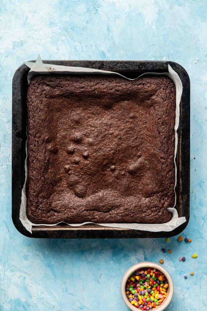 A square Cosmic Brownies chocolate cake in a baking tray lined with parchment paper sits on a blue surface, with a small bowl of colorful sprinkles beside it.