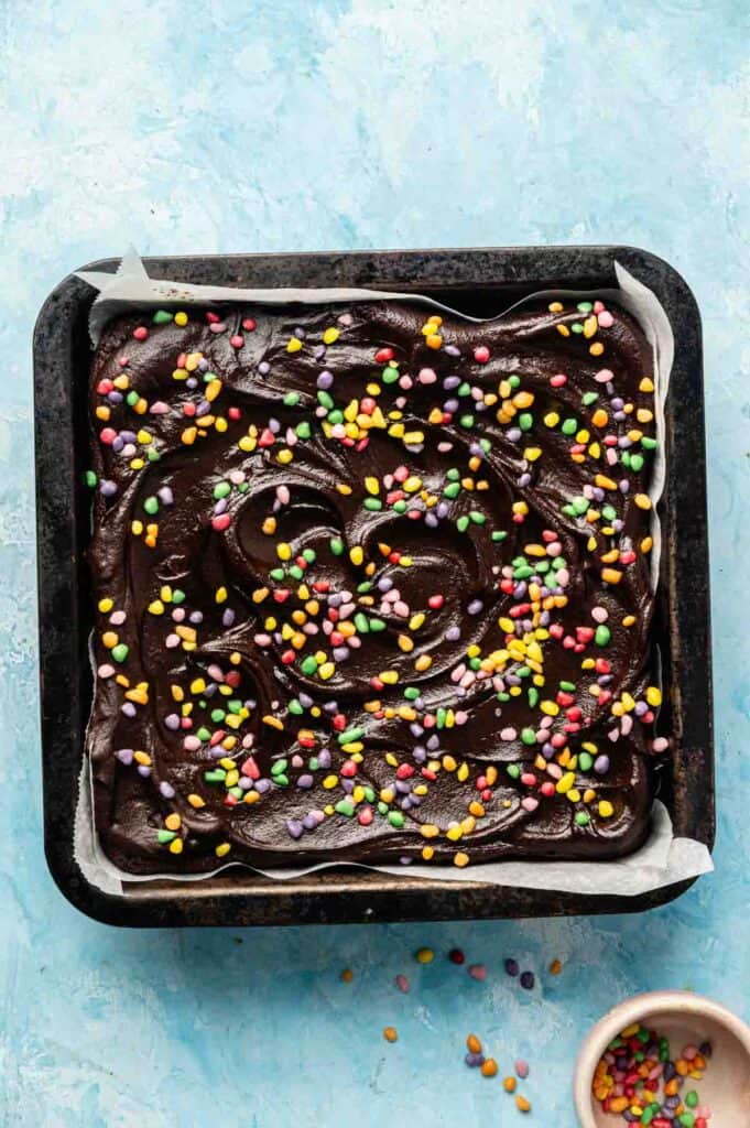 A square pan of Cosmic Brownies, chocolate frosted and topped with colorful round sprinkles, viewed from above on a light blue surface.