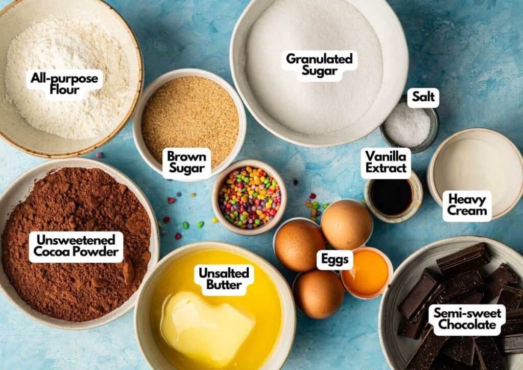A variety of baking ingredients in bowls for homemade Cosmic Brownies: flour, brown sugar, granulated sugar, salt, cocoa powder, vanilla extract, heavy cream, butter, eggs, mini candies, and semi-sweet chocolate.