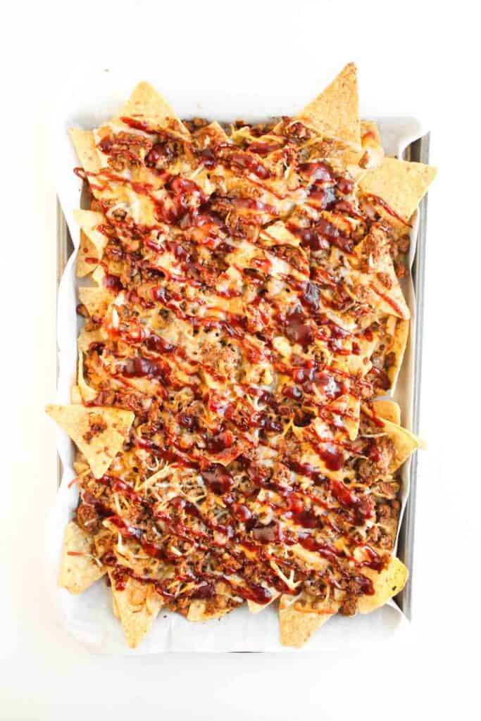 A baking tray filled with Cowboy Nachos topped with ground meat, melted cheese, and drizzled with barbecue sauce.