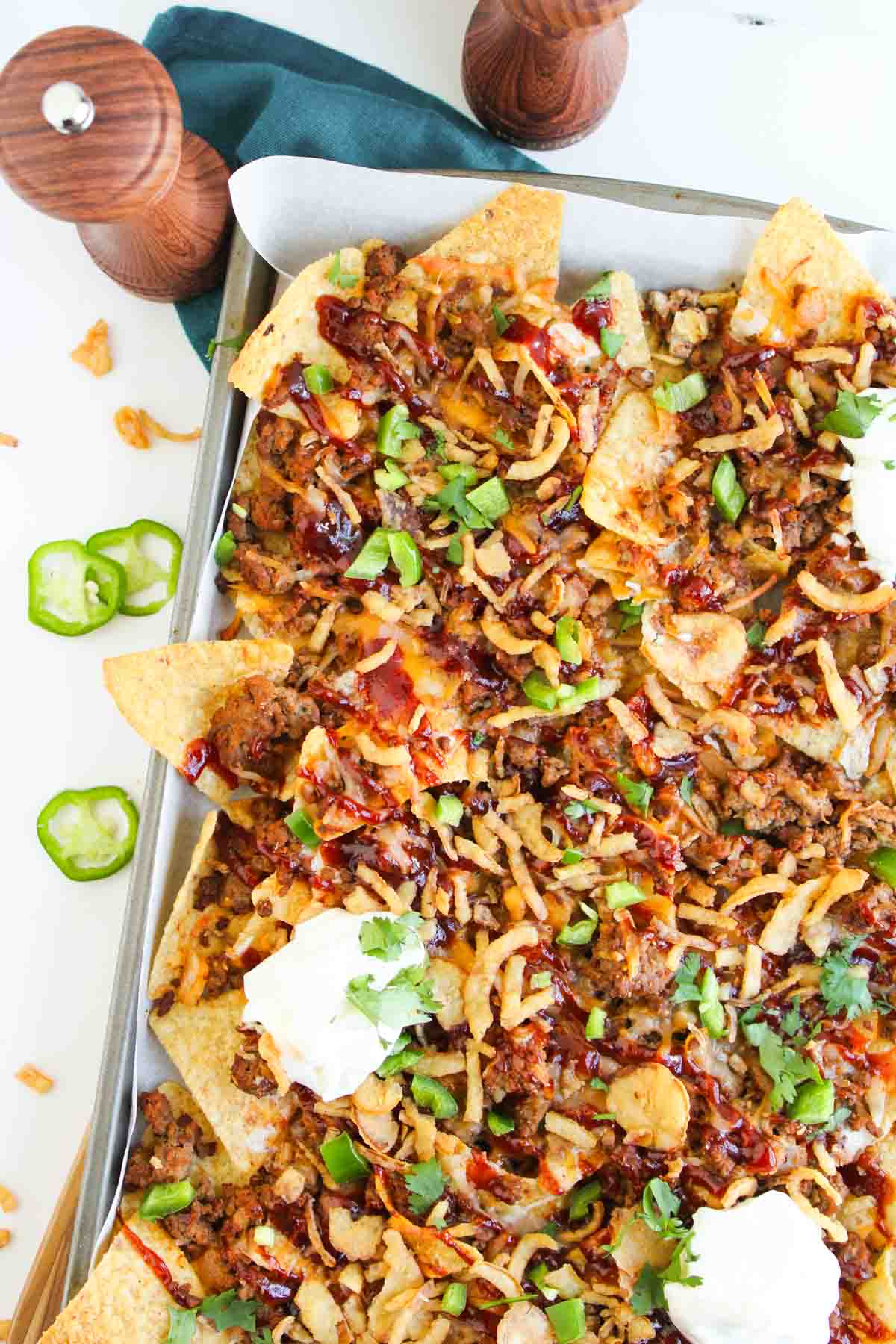 Cowboy Nachos: Sheet pan nachos topped with ground meat, melted cheese, barbecue sauce, sour cream, sliced jalape&ntilde;os, and chopped cilantro, with salt and pepper shakers in the background.