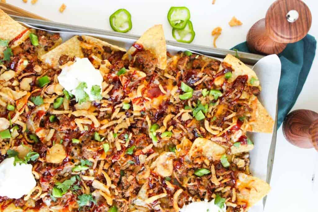 A tray of Cowboy Nachos topped with ground meat, crispy onions, cilantro, sliced jalape&ntilde;os, sour cream, and sauce sits ready to share, with salt and pepper shakers nearby.