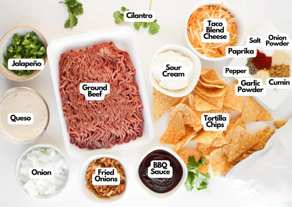 Ingredients for Cowboy Nachos are laid out, including ground beef, tortilla chips, queso, sour cream, BBQ sauce, spices, jalape&ntilde;o, onion, fried onions, taco cheese, and cilantro.
