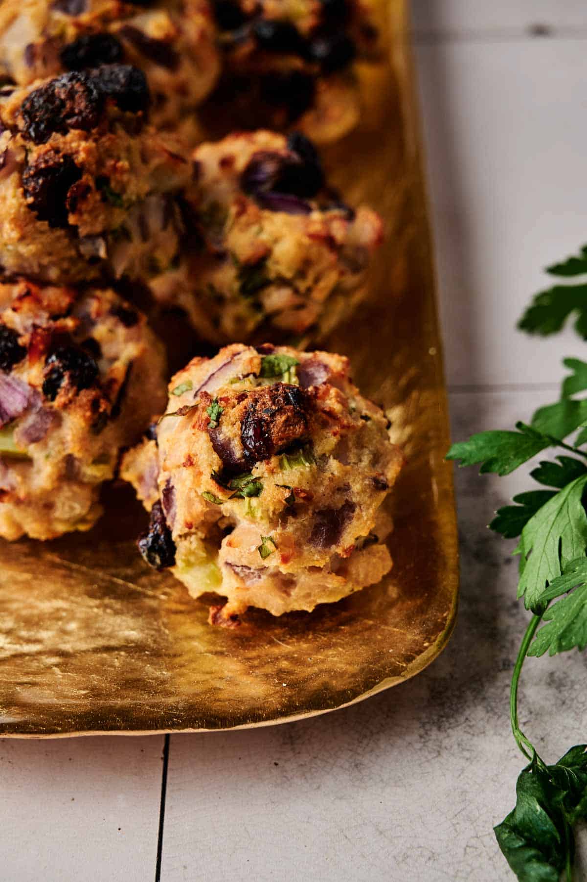 Close-up of golden-brown Cranberry Turkey Stuffing Balls with visible herbs and diced vegetables on a gold tray, garnished with fresh parsley leaves on the side.