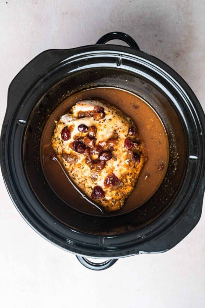 A cooked Crockpot Turkey Breast with sauce and cranberries sits in a black slow cooker on a light-colored surface.