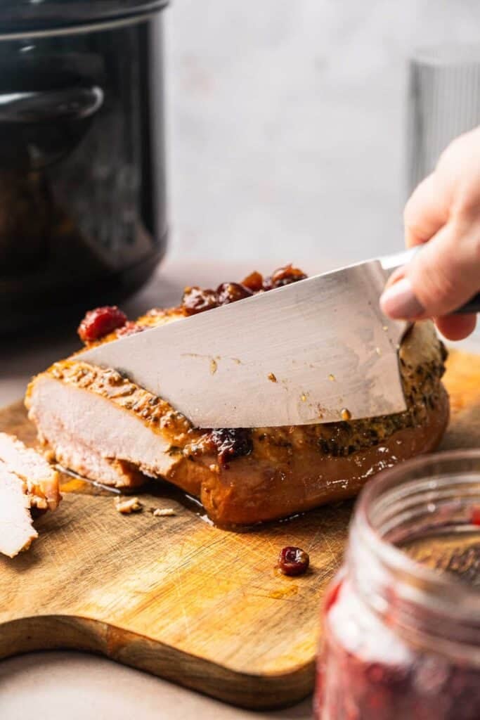 A hand slices Crockpot Turkey Breast on a wooden cutting board, topped with cranberries, while a jar of cranberry sauce sits nearby.