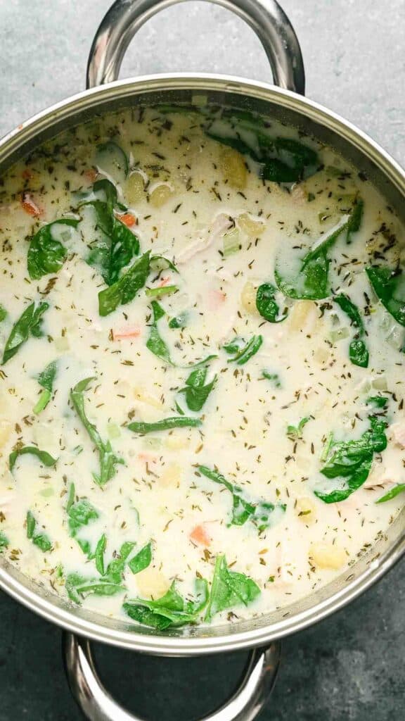 A pot of creamy Gnocchi Soup with visible spinach leaves, herbs, carrots, and pieces of chicken sits on a gray surface.