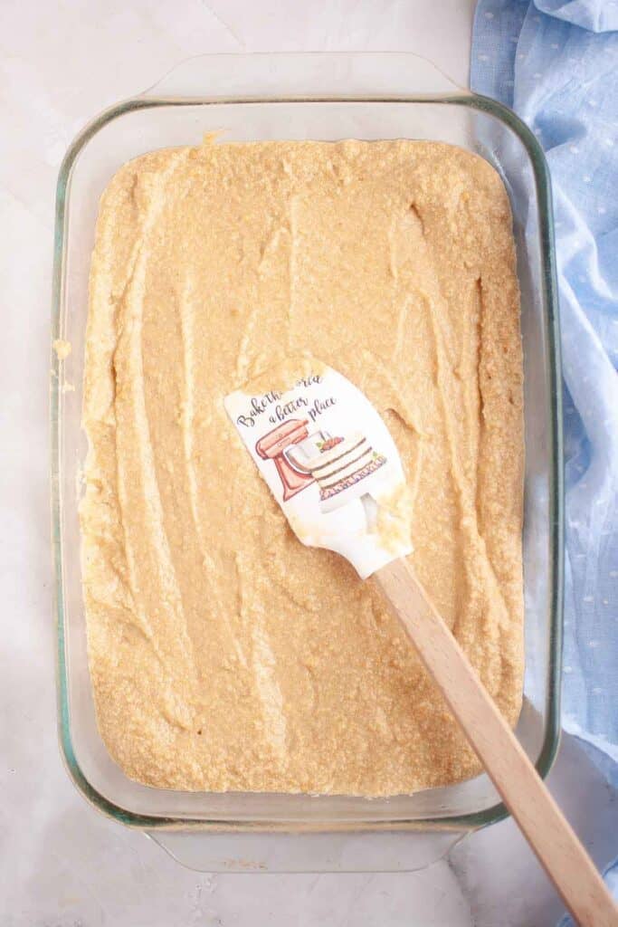 A glass baking dish filled with an even layer of light-colored Graham Cracker Cake batter, with a spatula resting on top.