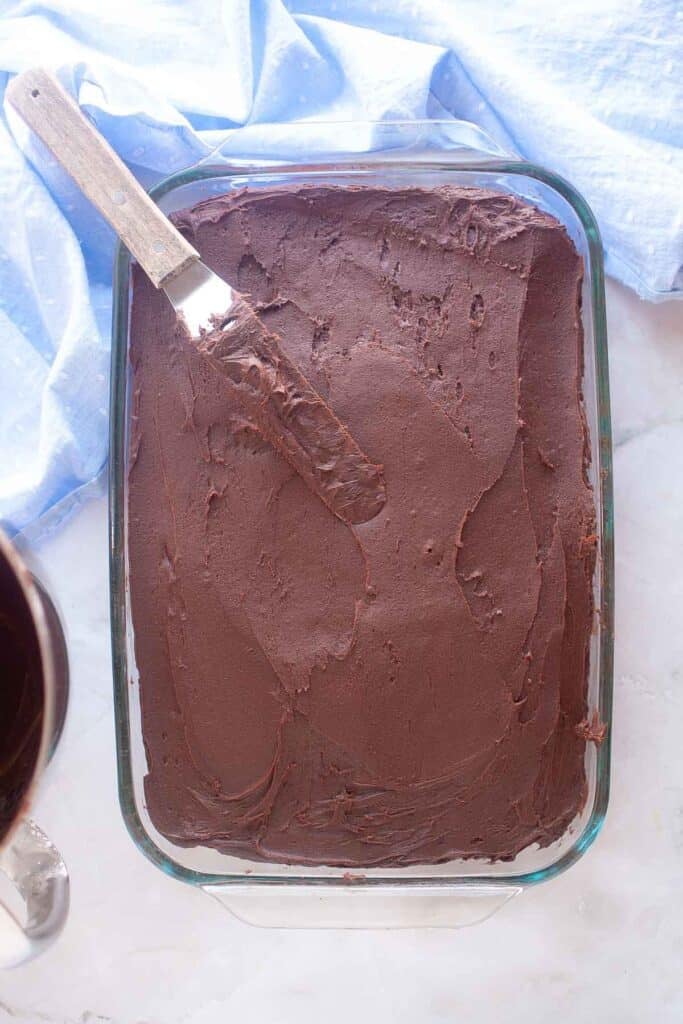 A rectangular glass baking dish filled with chocolate-frosted Graham Cracker Cake sits on a marble surface, with a spatula resting on top.