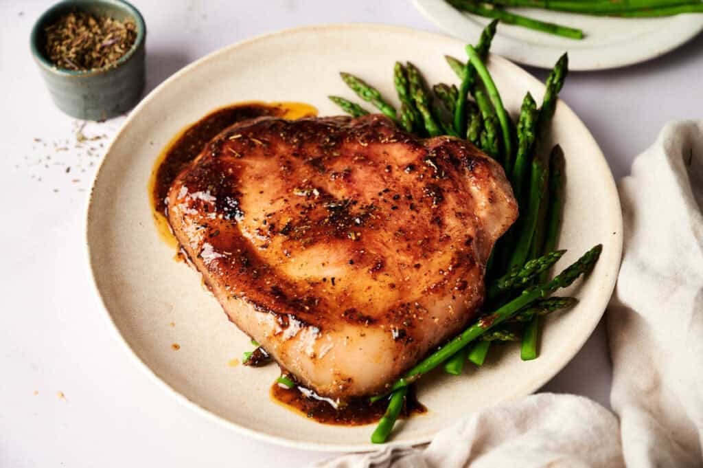 A cooked pork chop or ham steak topped with brown sauce sits on a plate beside steamed asparagus, accompanied by a small bowl of seasoning.