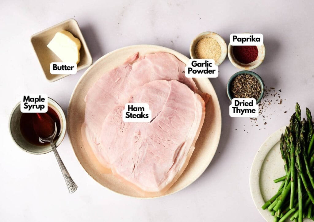 A plate of ham steaks is surrounded by small bowls of butter, maple syrup, garlic powder, paprika, dried thyme, and a side of asparagus, all arranged on a white surface.