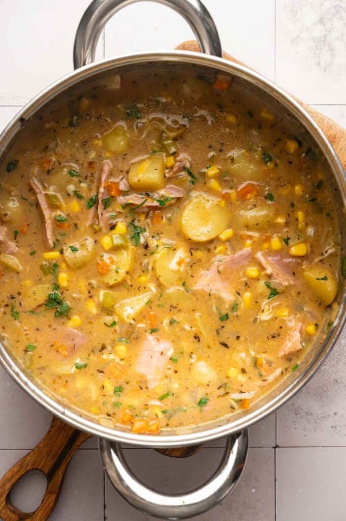 A pot of creamy Ham and Potato Soup, filled with chunks of potato, corn, shredded ham, carrots, herbs, and broth, sits on a wooden trivet.