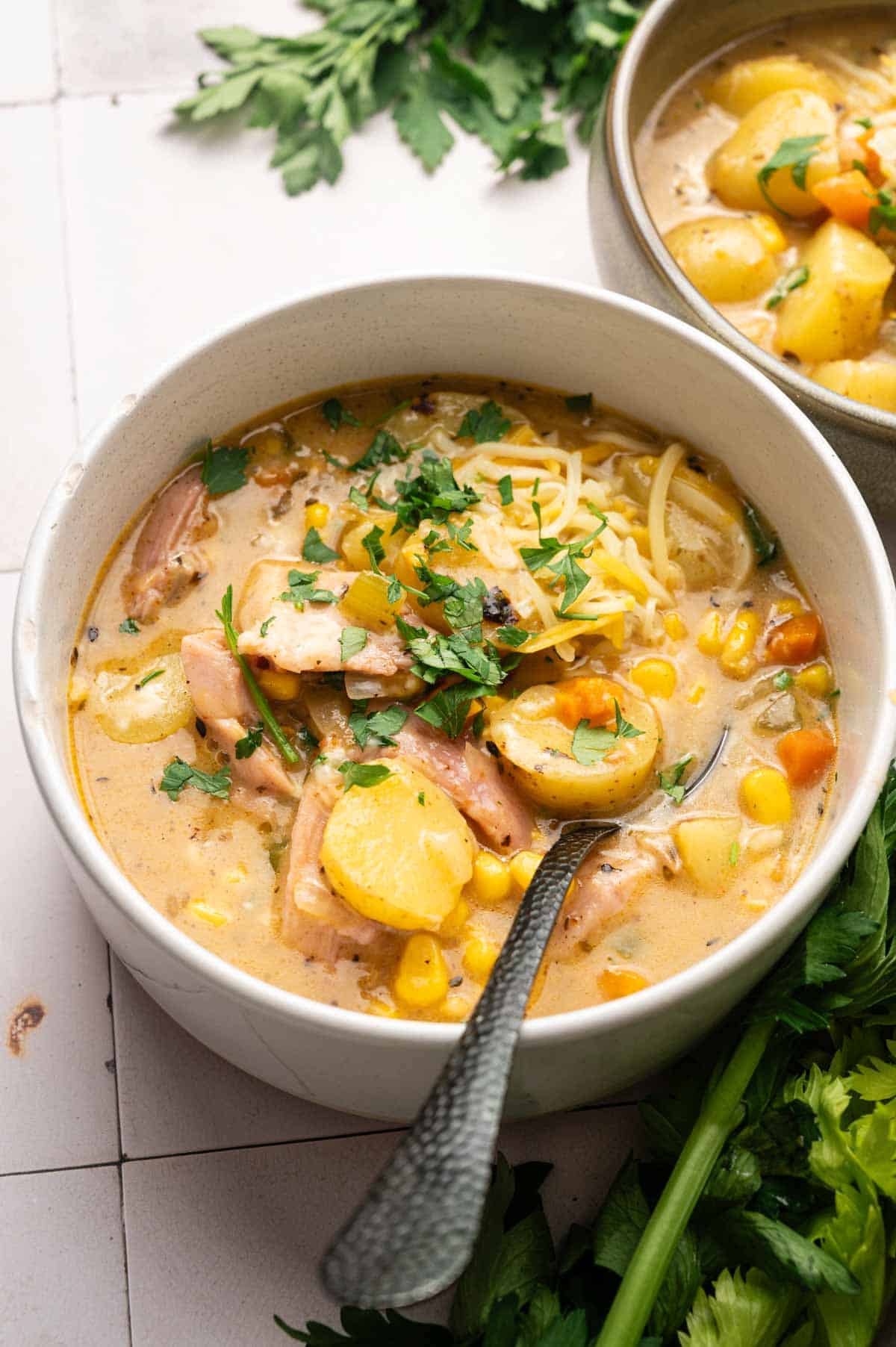 A bowl of creamy Ham and Potato Soup with chicken, corn, carrots, shredded cheese, and fresh herbs sits ready with a spoon. Another bowl of soup and sprigs of fresh herbs are nearby for garnish.