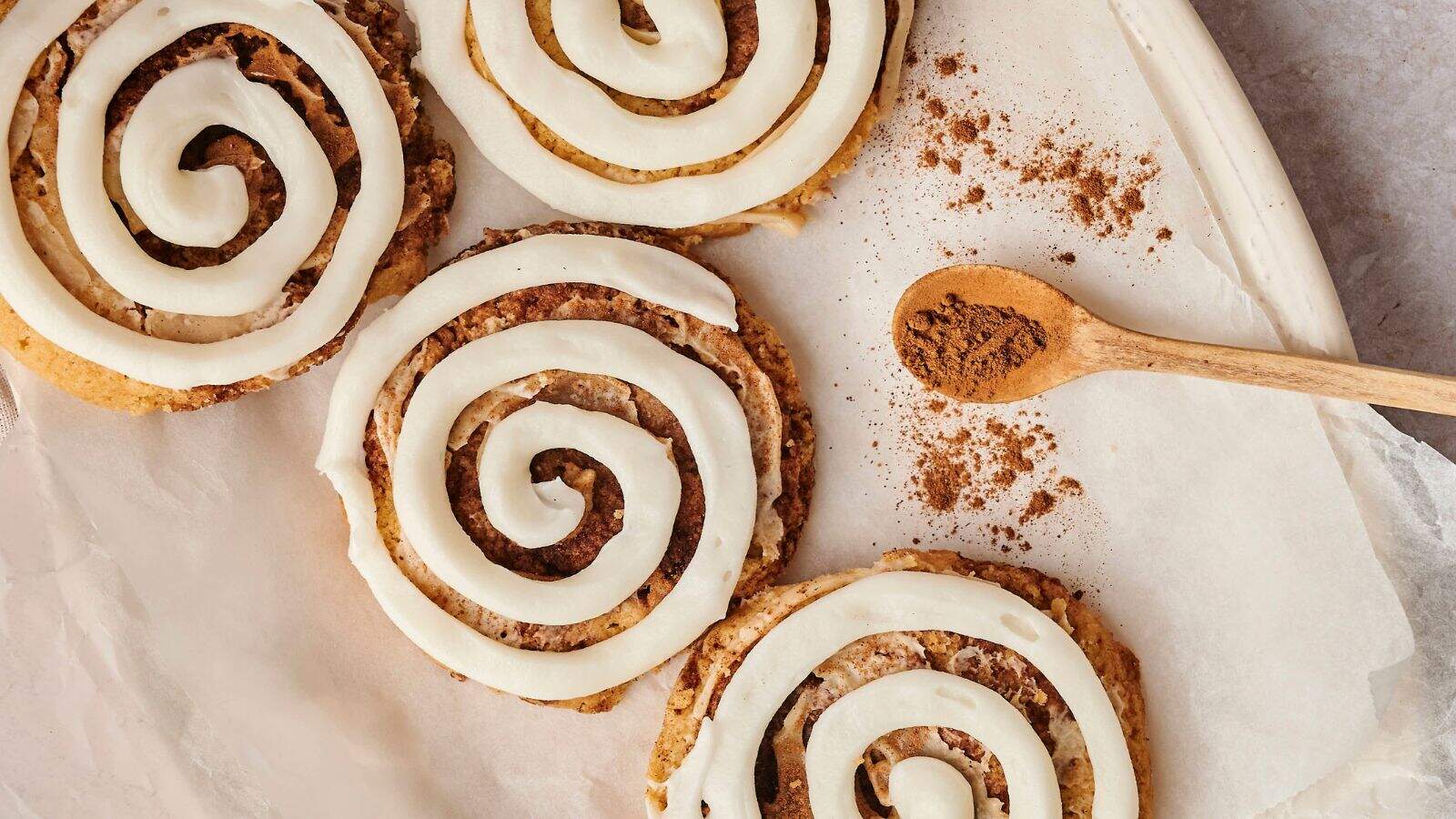 Four cinnamon rolls with white icing sit on parchment paper beside a wooden spoon containing ground cinnamon.