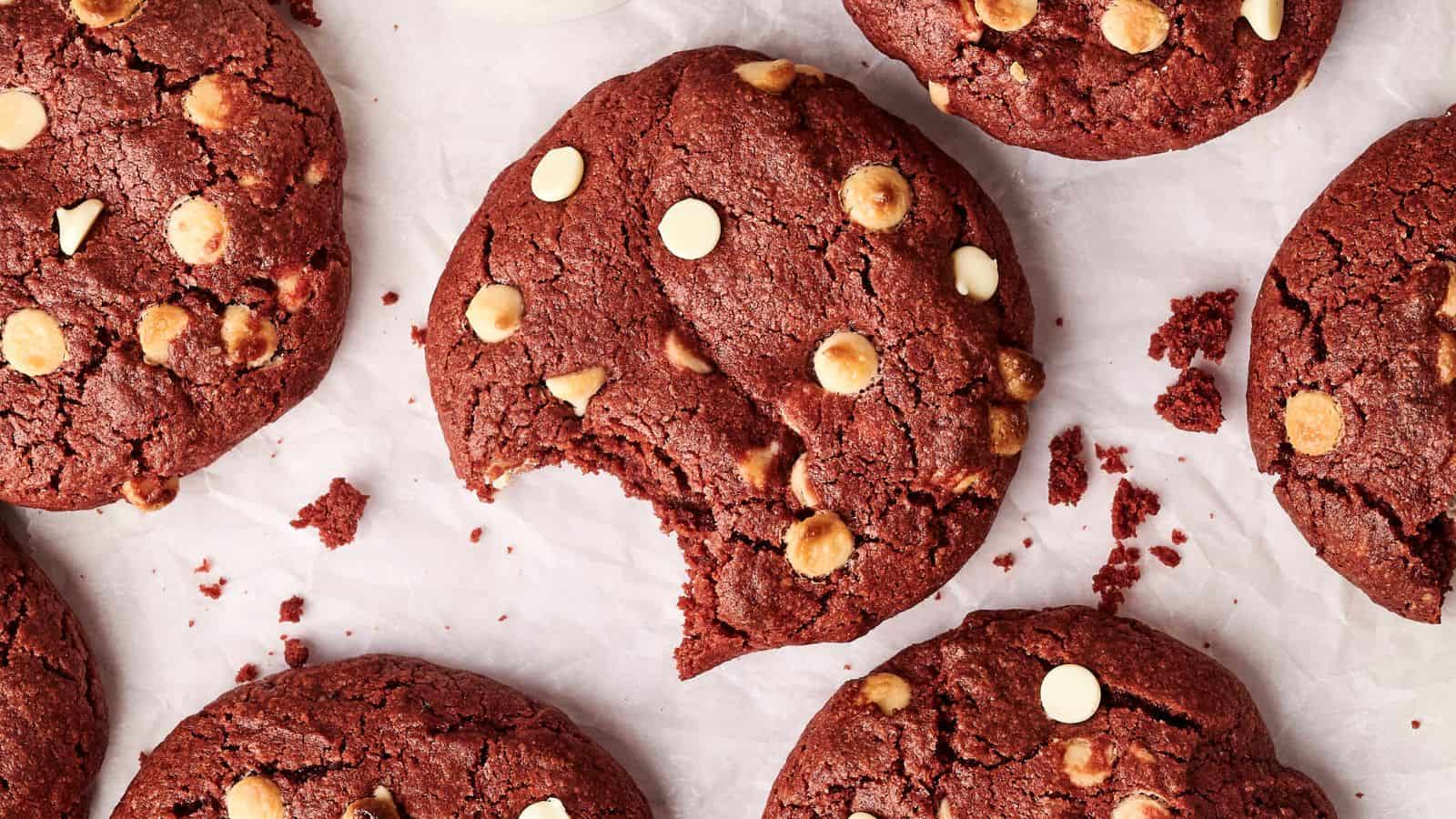 Several red velvet cookies with white chocolate chips on parchment paper, with one cookie in the center that has a bite taken out of it.