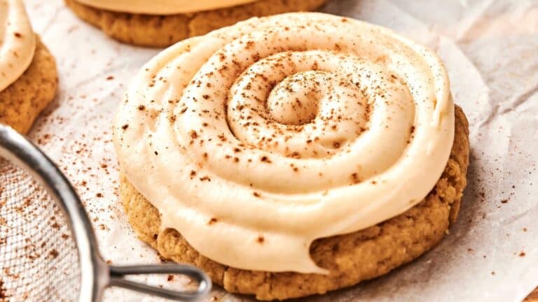 A frosted cookie with a spiral of creamy icing on top, sprinkled with a light dusting of cinnamon, resting on parchment paper.