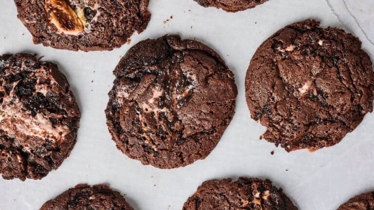 Seven chocolate cookies with a cracked surface and visible marshmallow pieces are arranged on a white parchment paper background.