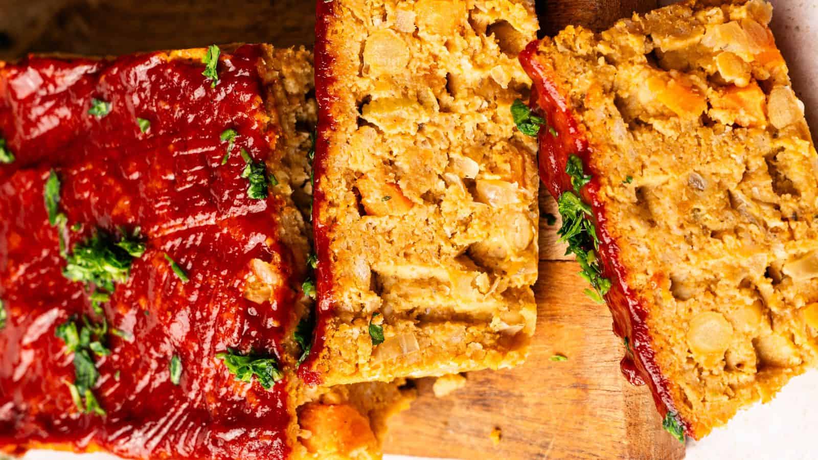 A close-up of sliced vegan meatloaf topped with ketchup and garnished with chopped parsley, displayed on a wooden cutting board.