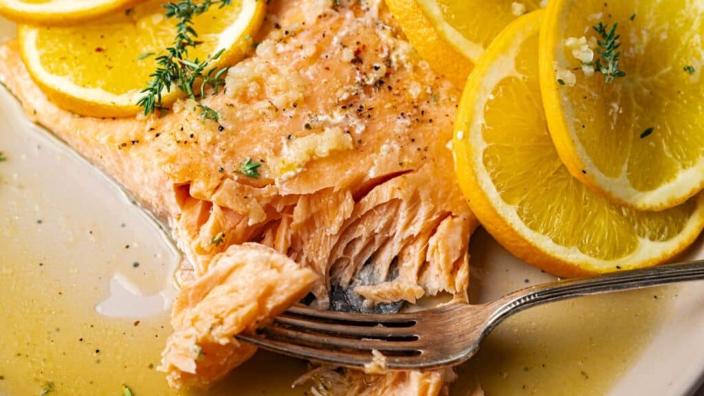 A close-up of cooked salmon garnished with lemon slices and herbs, with a fork flaking a piece of the fish on a plate with sauce.