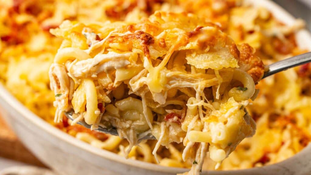 A close-up of a spoonful of cheesy baked pasta with shredded chicken and a golden, crispy topping, held over a dish filled with more of the same casserole.