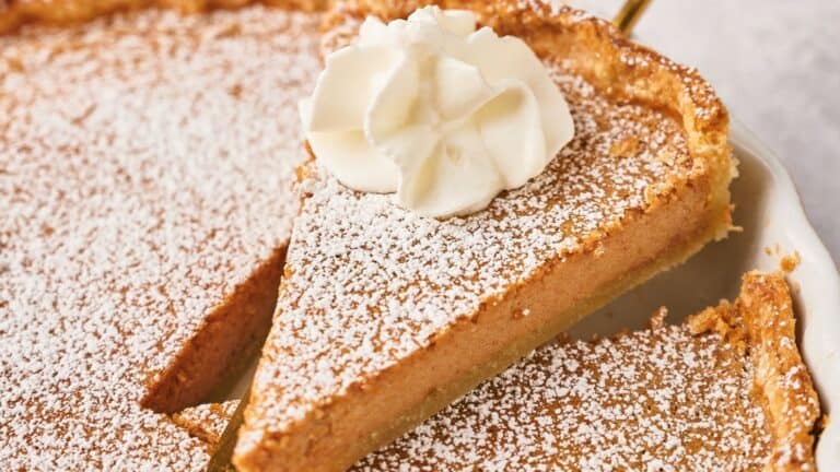 A close-up of a sliced custard pie topped with powdered sugar and a dollop of whipped cream on one slice.