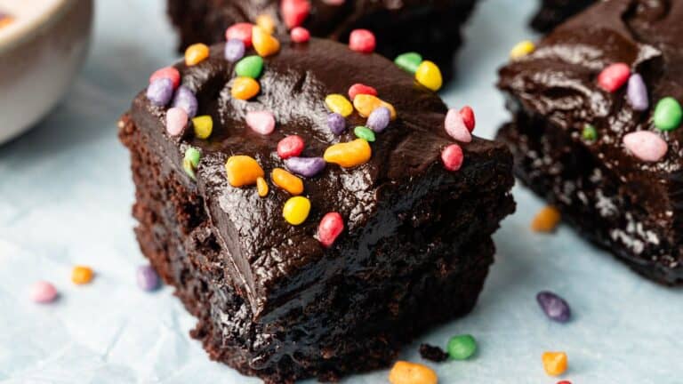A close-up of a chocolate brownie topped with colorful candy sprinkles, set on a light blue surface.