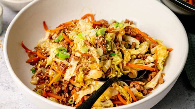 A white bowl filled with a stir-fry of ground meat, shredded cabbage, carrots, green onions, and sesame seeds, with a black spoon.