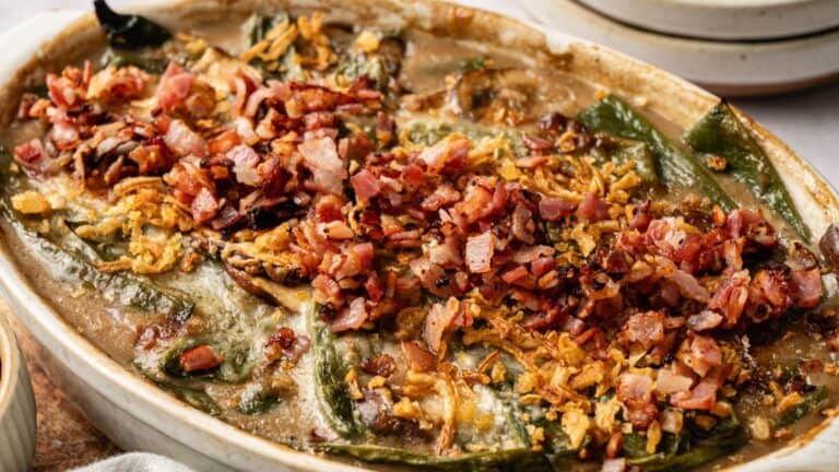 A casserole dish filled with stuffed poblano peppers, topped with crispy bacon bits and fried onions in a creamy sauce.