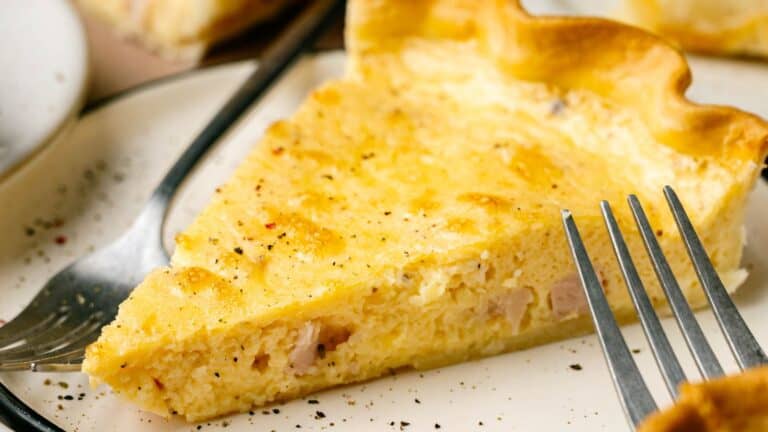 A slice of quiche on a white plate with a fork, showing a golden crust and creamy filling with visible seasoning.