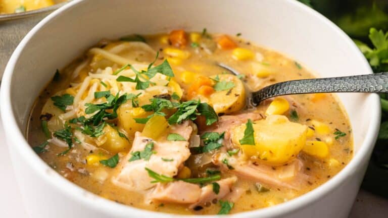 A bowl of hearty chicken and vegetable soup with potatoes, corn, carrots, herbs, and a spoon resting inside.