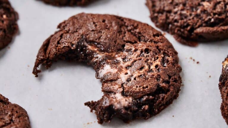 A chocolate cookie with a gooey center and a bite taken out, resting on white parchment paper. Other similar cookies are partially visible around it.