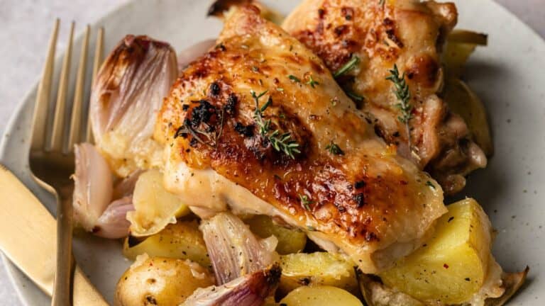 Roasted chicken thighs served on a plate with golden potatoes, caramelized onions, and garnished with fresh thyme.