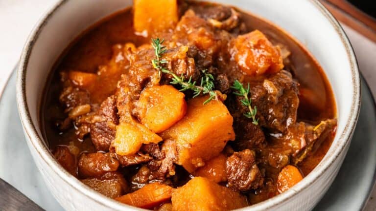 A bowl of beef stew with chunks of beef, carrots, potatoes, and a sprig of fresh herbs in a rich, brown broth.